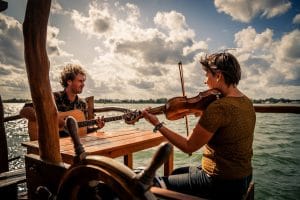 Florian und Anouk sitzen für ihr Album "Port de Taipana" mit Gitarre und Violine auf der sonnenbeschienenen Veranda ihres Hausbootes.
